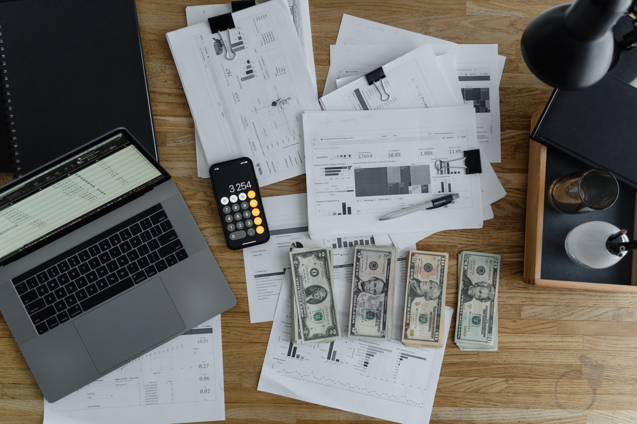 Dollar bills, an iPhone and a laptop on an office table next to piles of paper printed with graphs.