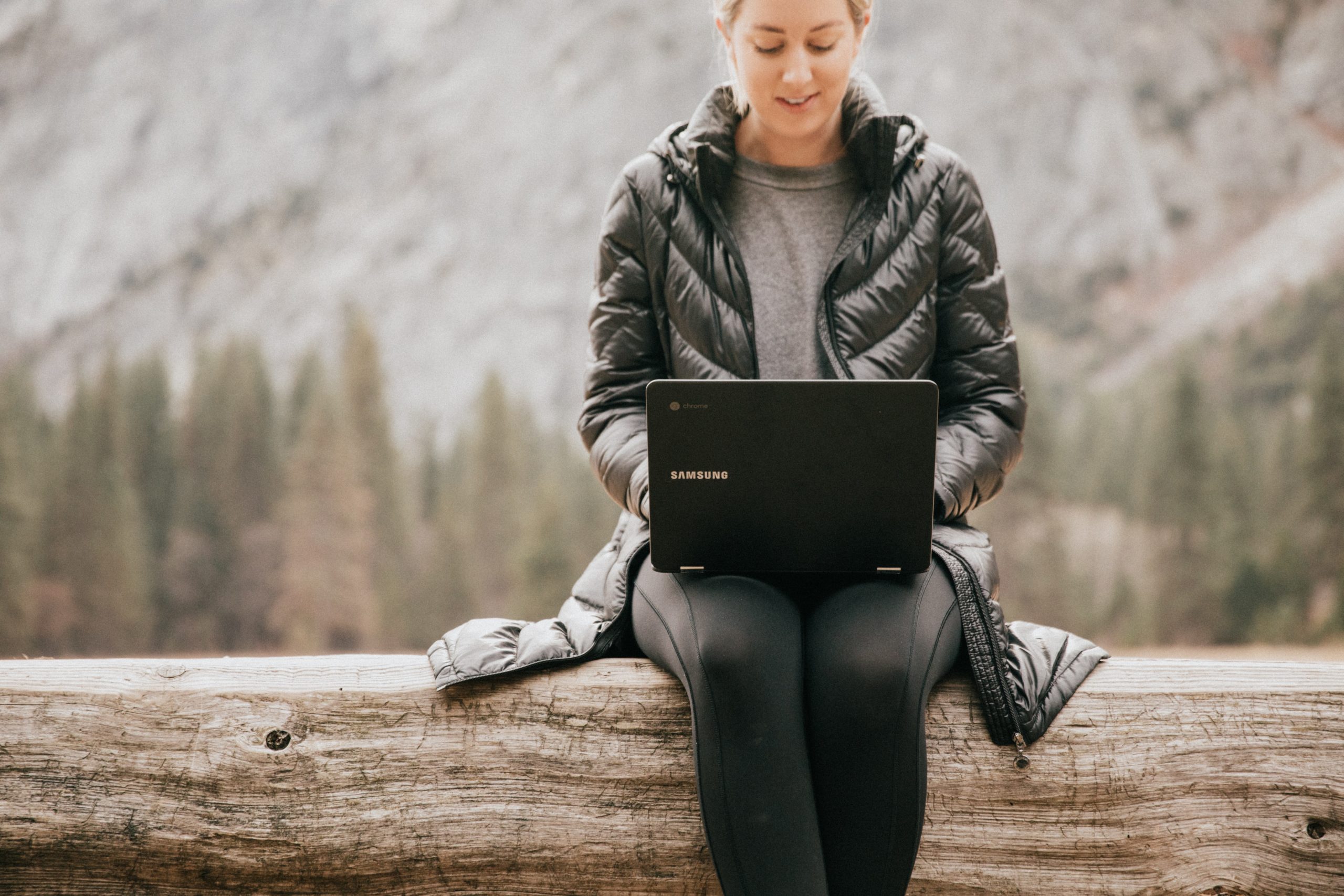 A woman working on a laptop outside sitting at a wooden log.