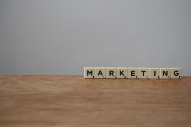 The word “marketing” is spelled out with Scrabble tiles on a brown wooden table.