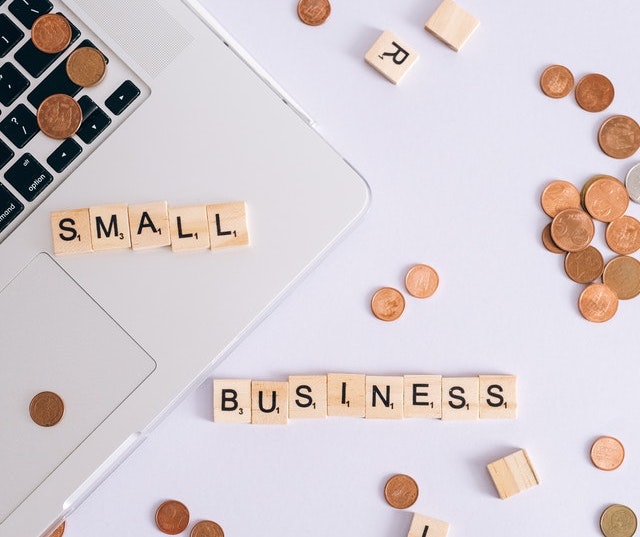 The term “small business” is spelled out with Scrabble tiles on a table alongside coins and a laptop