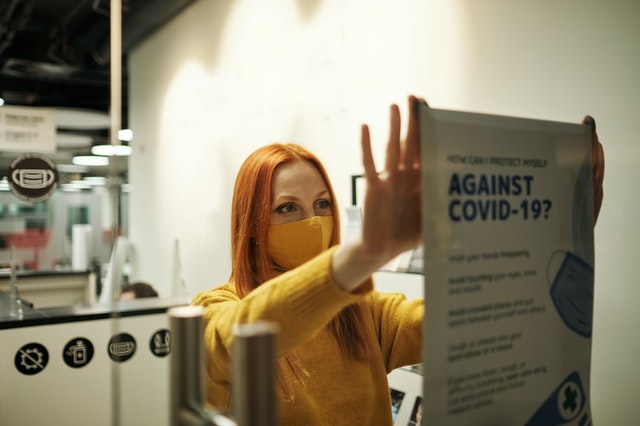 Woman wearing a mask holds a white piece of printer paper that says “against COVID.”