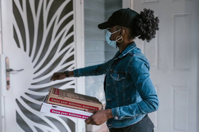 A delivery woman standing at the door wearing a mask holding a pizza