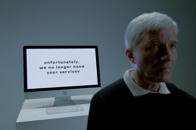 Man seated next to a screen that says, “unfortunately we no longer need your services.”