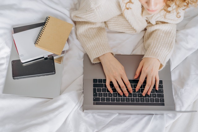 A woman in a sweater typing on a laptop with books kept aside while in bed
