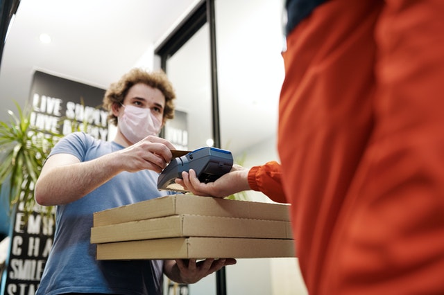 A man wearing a face mask paying for his pizza delivery through a billing machine.